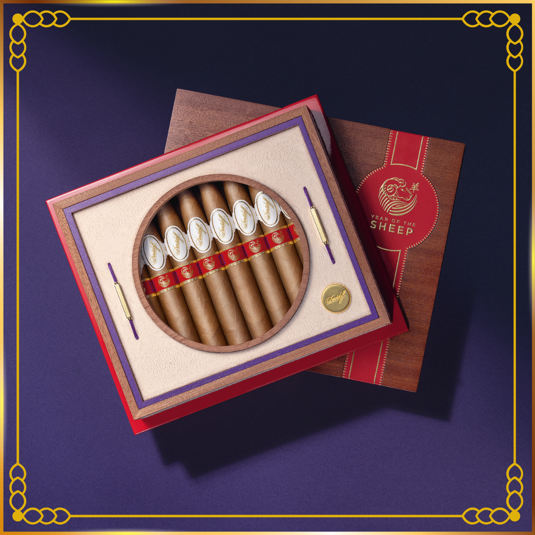 A premium Davidoff Year of the Sheep 2027 cigar box featuring an open red-and-cream presentation box with a circular window displaying several brown cigars wrapped in a red band, alongside a closed brown box with a red ribbon and a gold crest. The set is photographed on a dark purple background with a decorative gold border.
