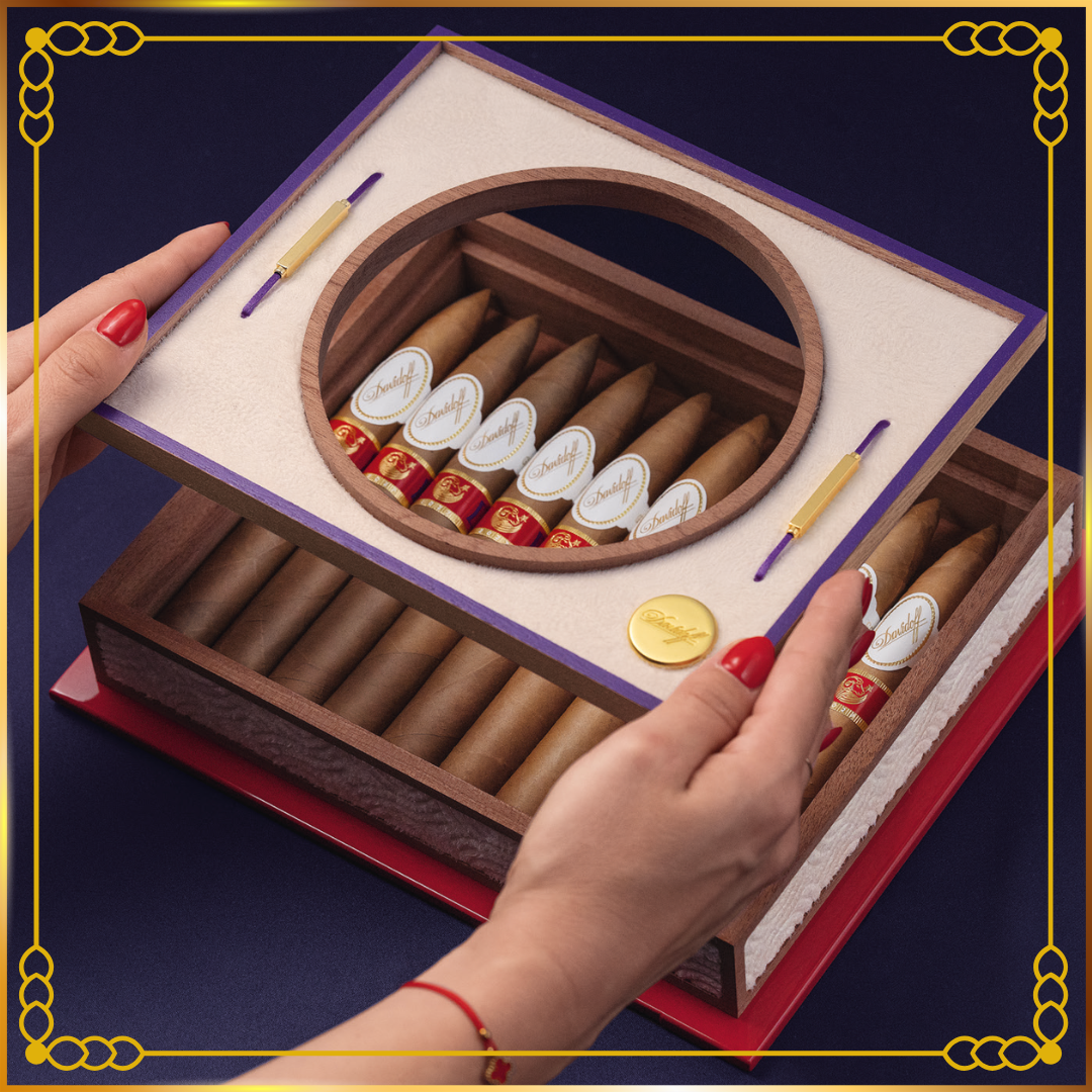 A luxury Davidoff Year of the Sheep 2027 cigar box with a hinged lid, shown with hands lifting the lid. The box contains multiple cigars with white-and-red bands, a gold coin, and two decorative gold-and-purple tools, all on a dark blue background.