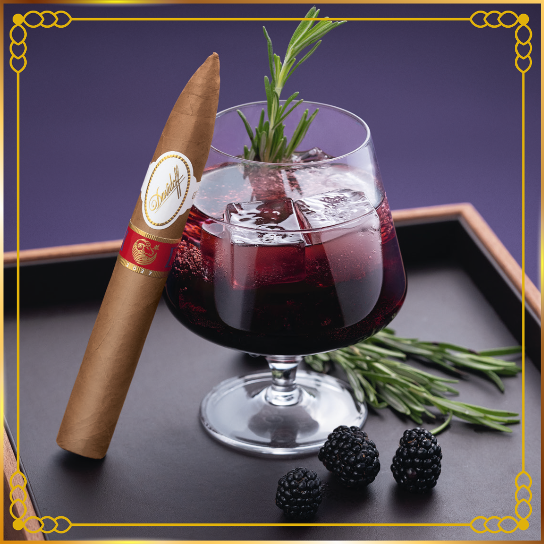A Davidoff Year of the Sheep 2027 cigar and a glass of dark red cocktail with rosemary garnish, styled on a tray with blackberries against a purple background.