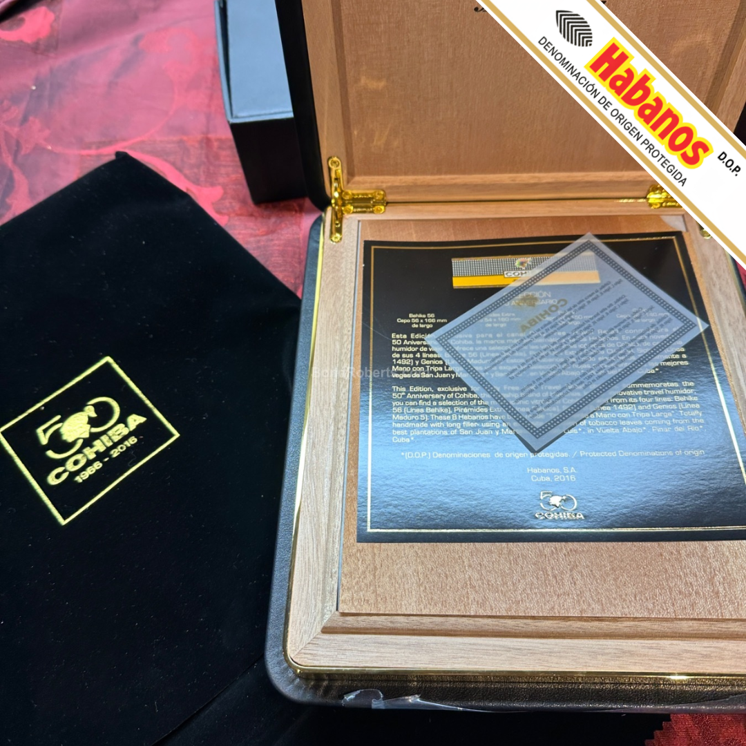 A wooden Cohiba Selección 50th Anniversary Travel Humidor opened to display a commemorative overview of the cigars and certificate, alongside a black velvet Cohiba pouch with a gold logo and a Habanos banner tag on a red textured surface.