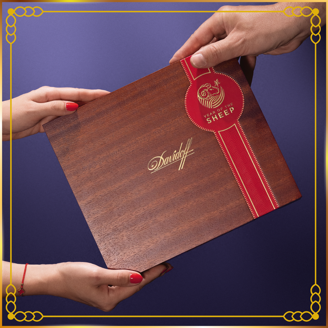 A wooden Davidoff Year of the Sheep 2027 presentation box with a red ribbon seal and gold script logo, held by four hands against a dark blue background with a gold decorative border.