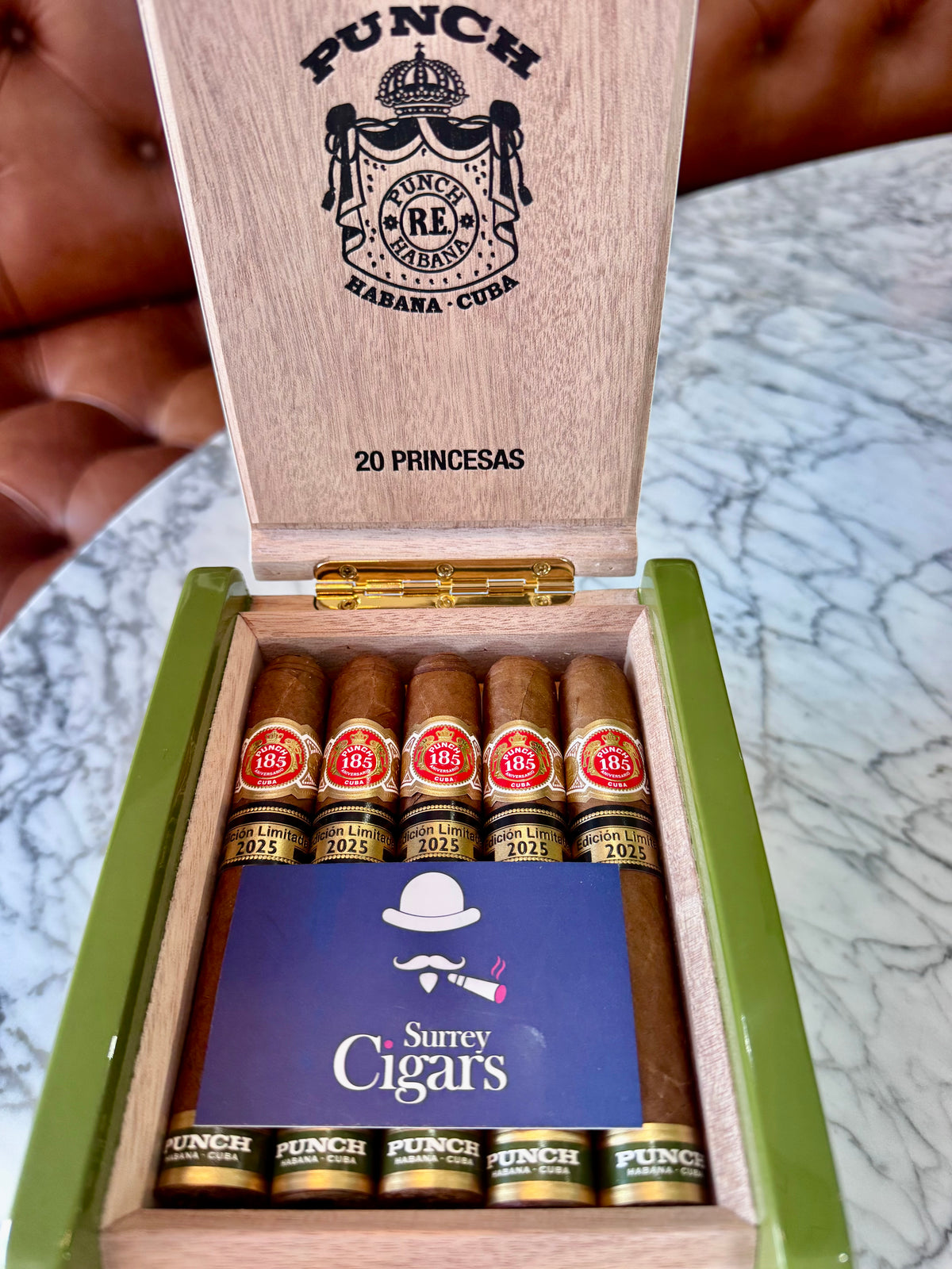 An open wooden cigar box branded PUNCH cigars, showing five Punch Princesas Limited Edition 2025 cigars with red-and-gold labels inside a green-lined box. A blue card reading 'Surreyy Cigars' is placed in front, and the box lid is open against a marble table background.