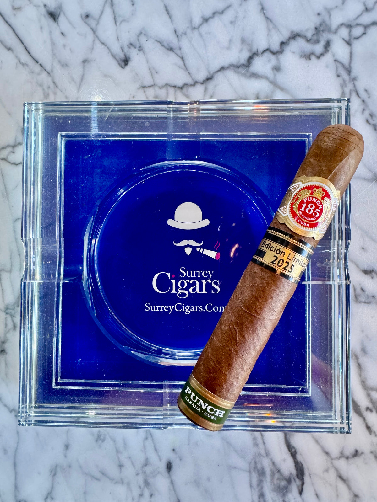 A single Punch Princesas Limited Edition 2025 cigar rests diagonally across a clear ashtray with a blue insert. The ashtray features the white 'Surrey Cigars' logo.